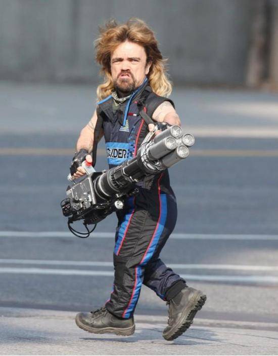 Peter Dinklage with a mullet, wearing a sleeveless jumpsuit, and holding a four-barrel laser cannon.